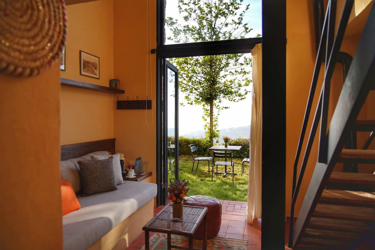 Ein gemütliches Zimmer mit einem Bett, einem kleinen Tisch mit einer Vase und Blick auf eine Rasenfläche und einen Baum durch eine geöffnete Tür.