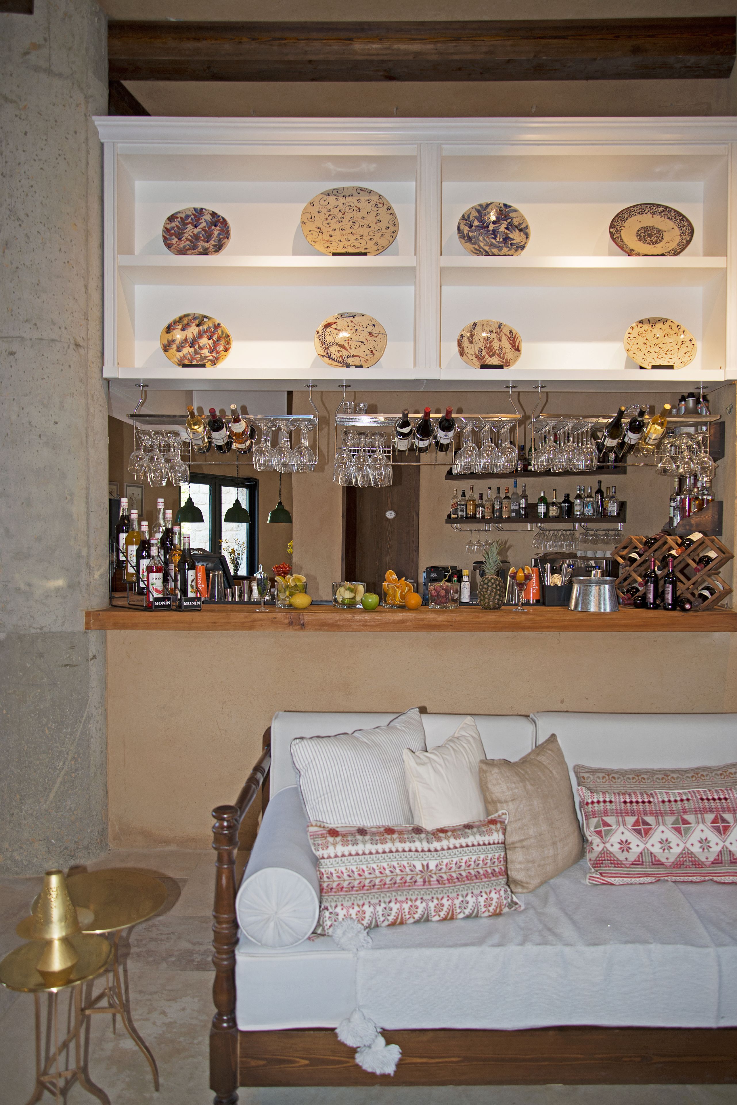 Un área de bar con estantes llenos de platos decorativos, diferentes botellas, copas y una barra de madera.