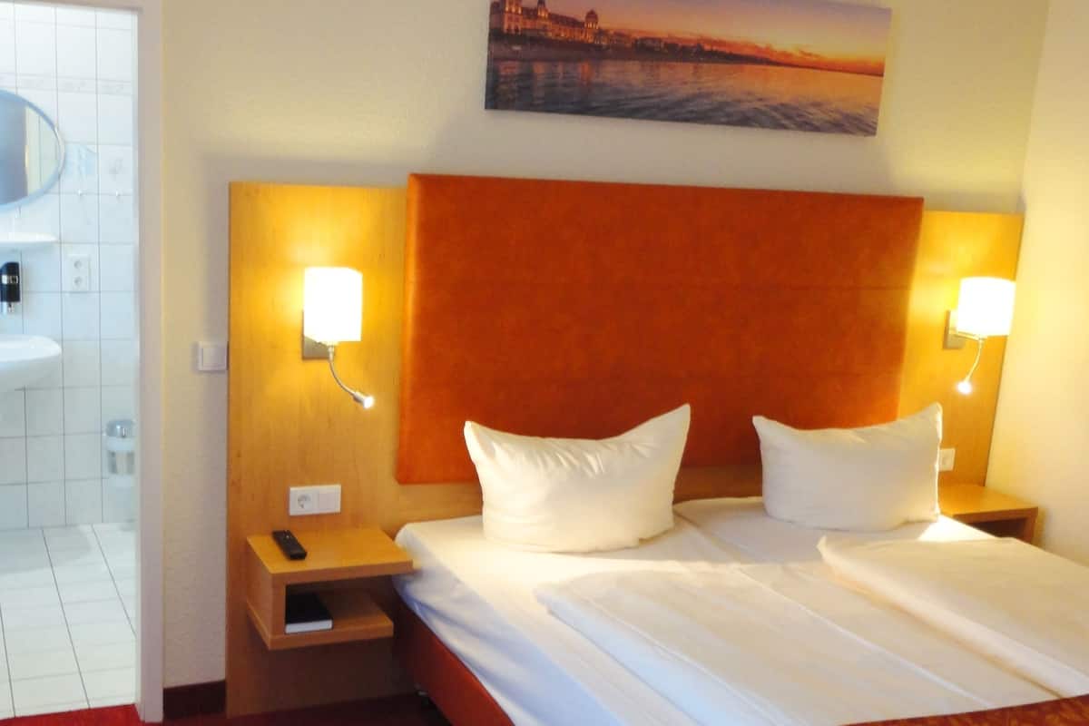 Una habitación de hotel con una cama grande, dos mesitas de noche, un velador, un cuadro en la pared y un baño visible a través de una puerta abierta.