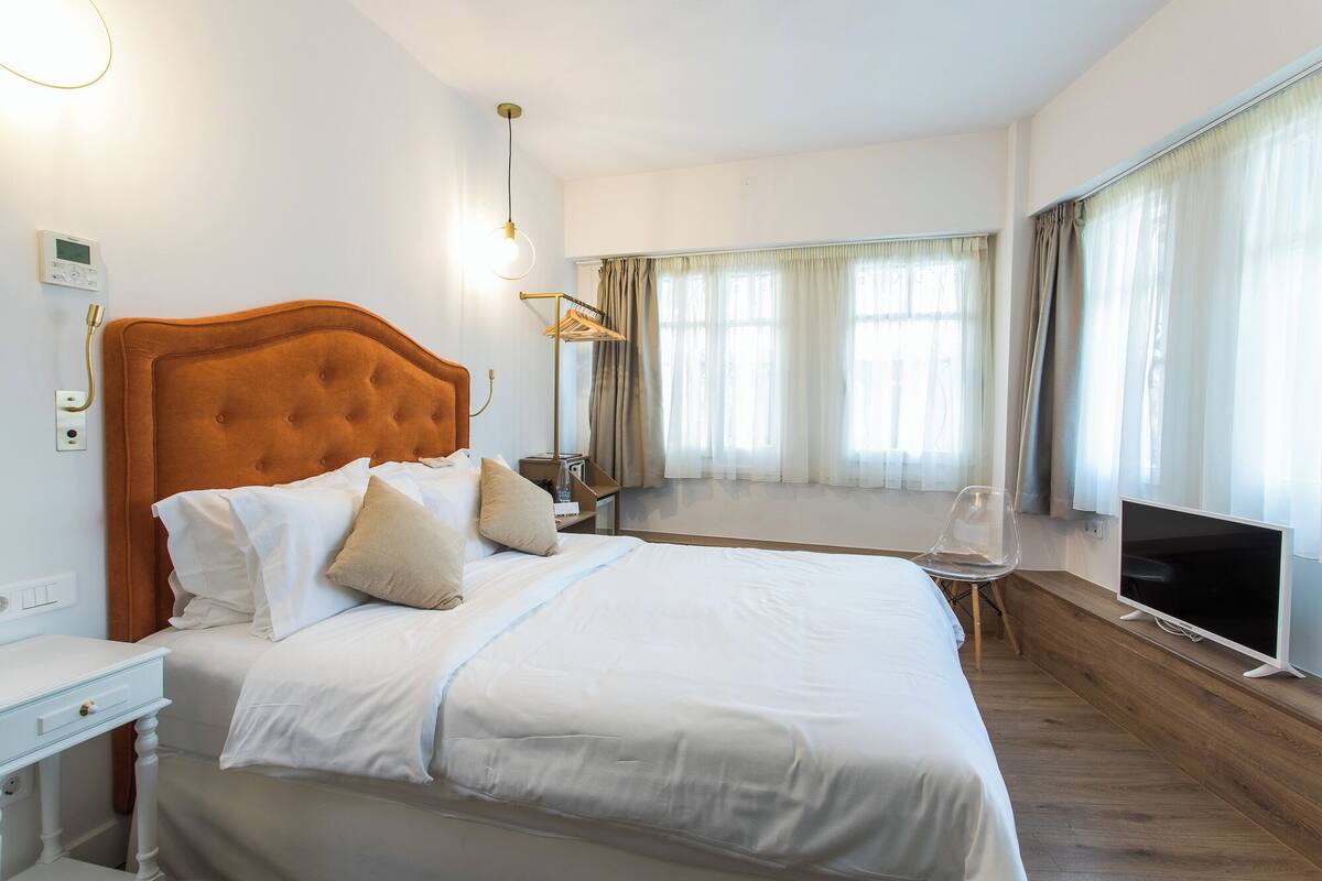 Habitación de hotel con cama, televisor, mesita de noche, calefactor, ventana con cortinas y termostato en la pared.