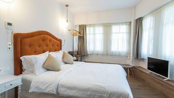 Une chambre d’hôtel équipée d’un lit, d’une télévision, d’une table de chevet, d’un radiateur, d’une fenêtre avec des rideaux et d’un thermostat fixé au mur.