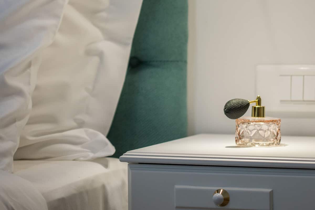 Una cama bien hecha con ropa blanca, un cabecero tapizado en verde, una mesita de noche blanca con un cajón y una lámpara fijada a la pared.