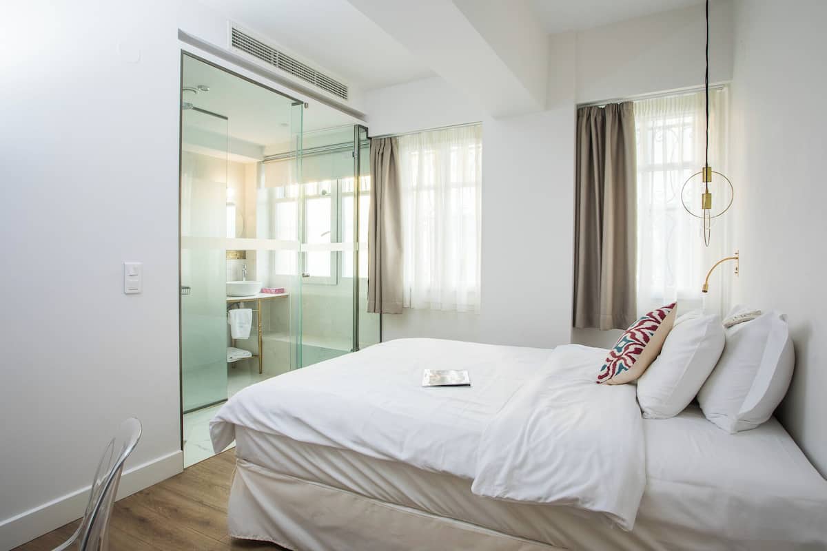 Un dormitorio con cama, mesita de noche, cabina de ducha de vidrio, ventana con cortinas y una lámpara.