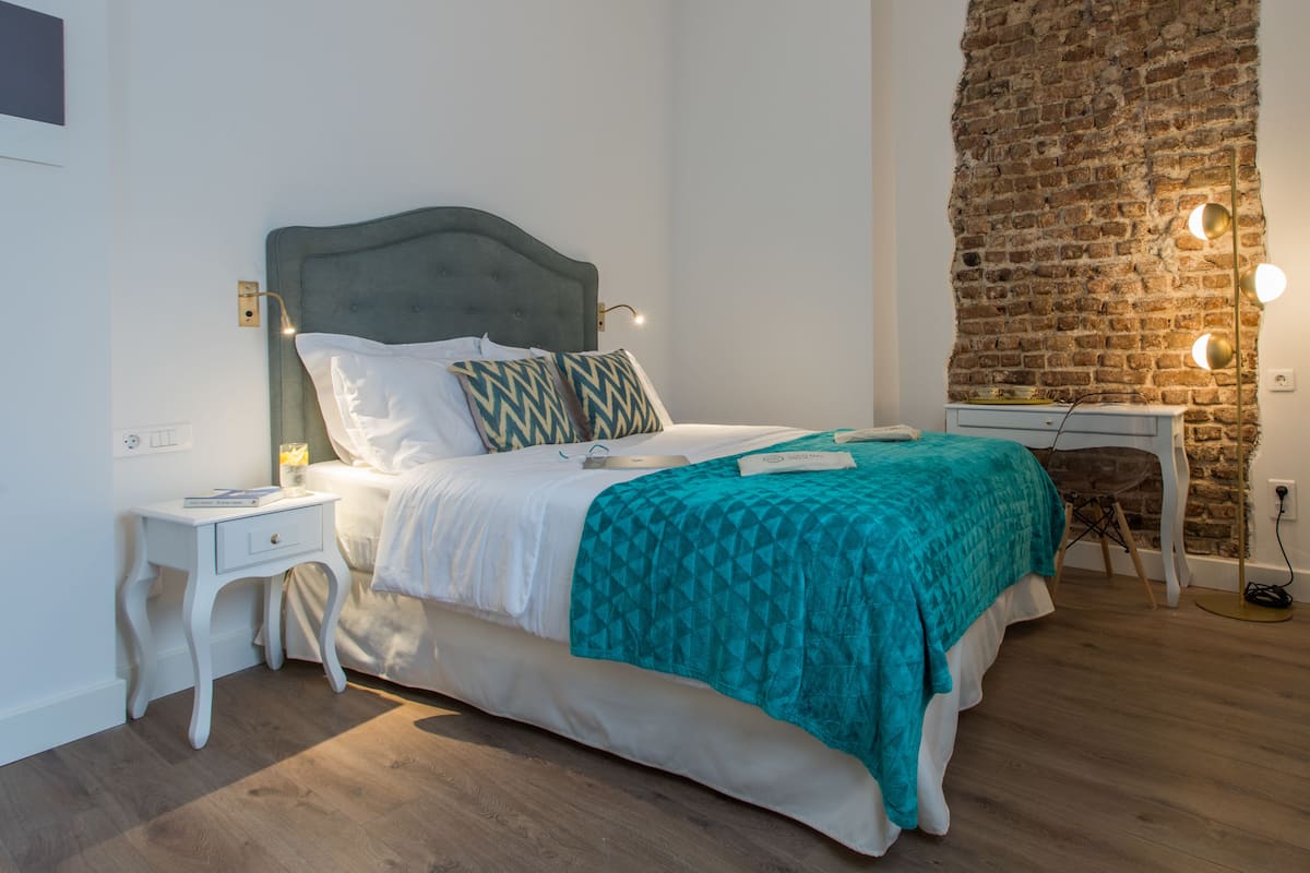 Una cama bien hecha con un edredón verde azulado, una almohada blanca y un cabecero gris. Se observa una mesita de noche blanca con una lámpara, y una pequeña mesa con una lámpara al lado de la cama. En el fondo, una pared de ladrillo.
