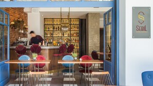 Un espace bar avec un barman, une étagère à bouteilles de vin et de spiritueux, et des fauteuils en velours rouge.