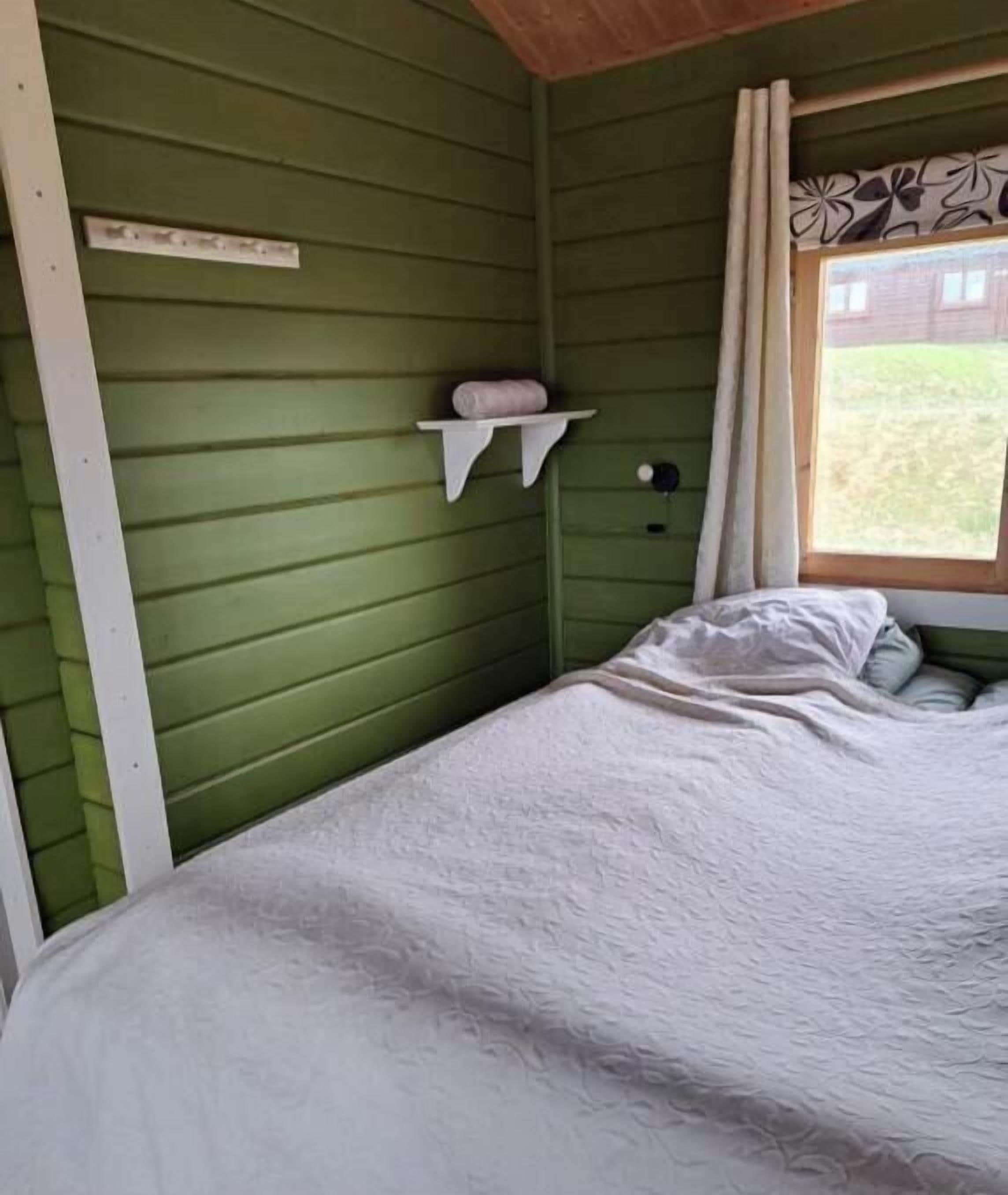 A room with a bed, a shelf with a pillow, a window with curtains, and a wall-mounted light.