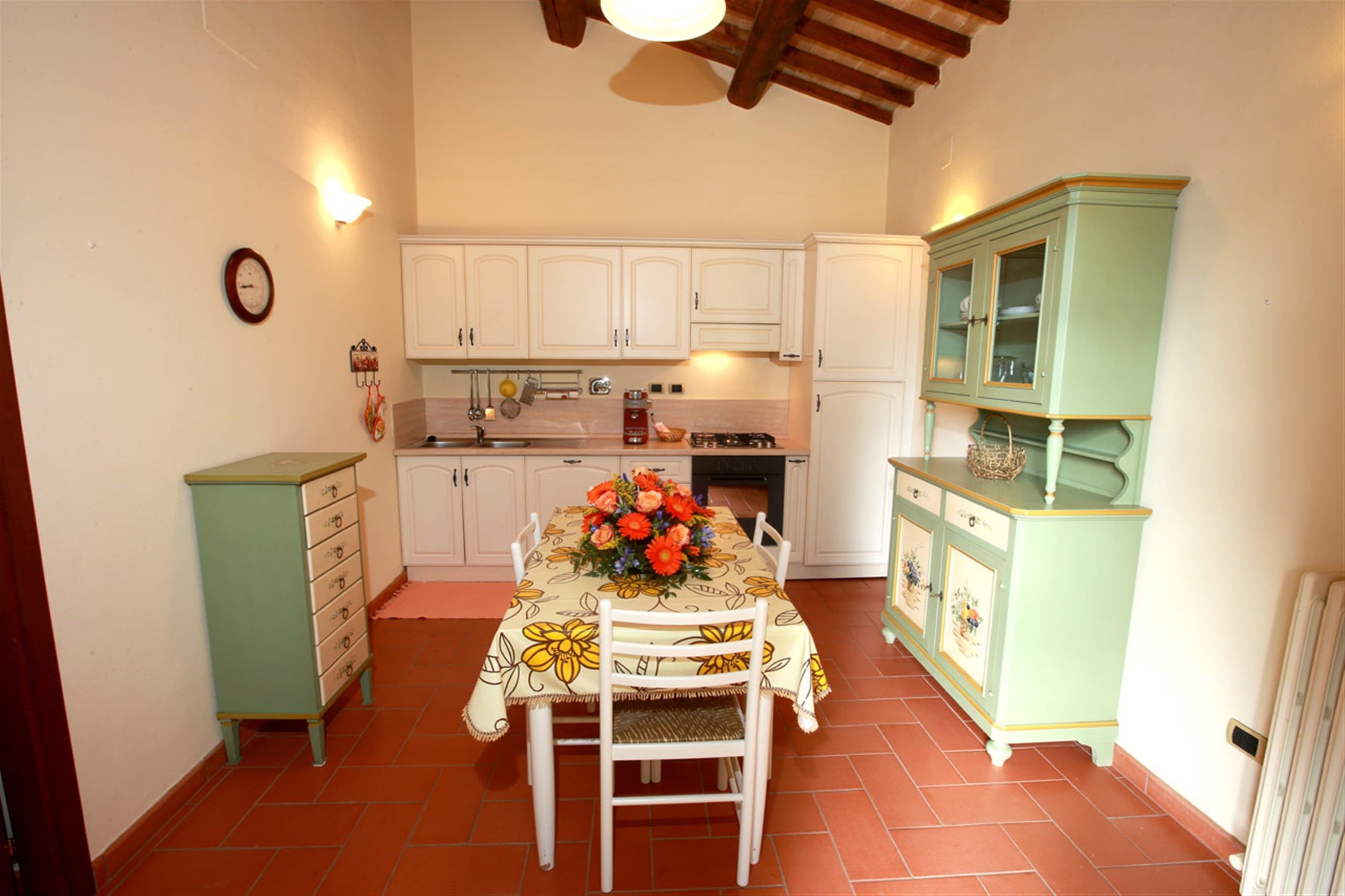 Una cocina con armarios blancos, una mesa de comedor con mantel floreado y un mueble verde con puertas de cristal.