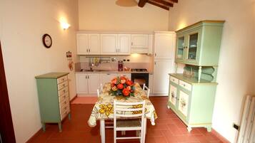 Una cocina con armarios blancos, una mesa de comedor con mantel floreado y un mueble verde con puertas de cristal.