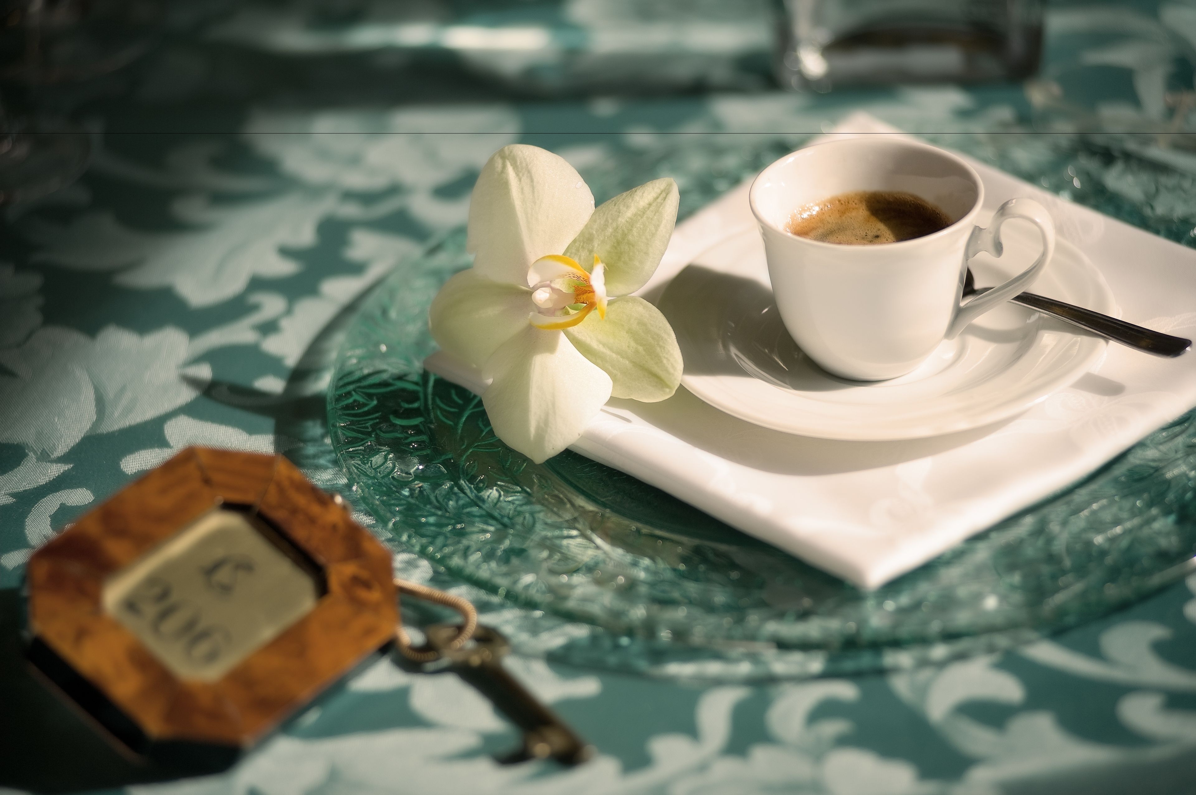 A white cup of coffee on a saucer with a spoon, a white orchid, and a keychain with a lock.