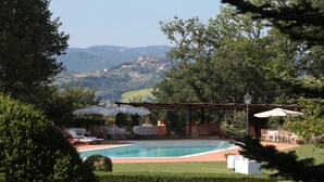 A pool area with lounge chairs and umbrellas, surrounded by greenery and a view of distant hills. - Il Castellaro Country House (Perugia)