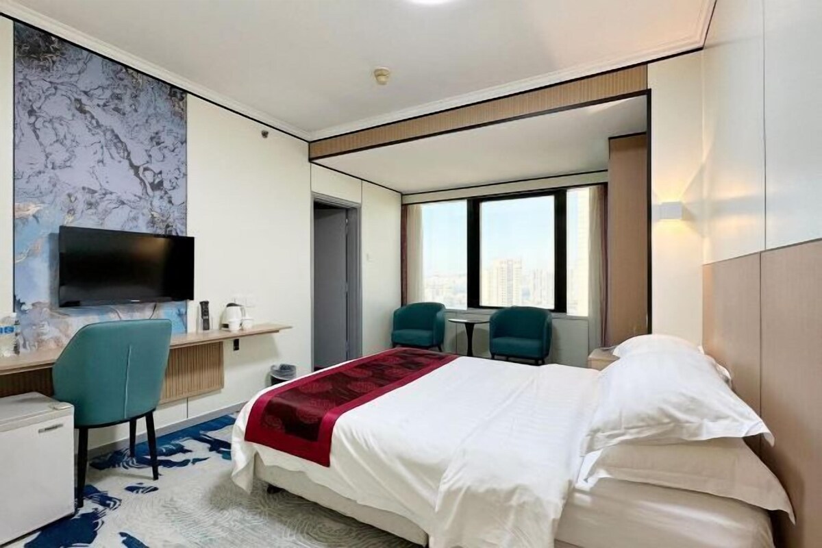 Een moderne hotelkamer met een bed, een bureau met een flatscreen-tv, twee stoelen, een bijzettafeltje en een groot raam met uitzicht op de stad.