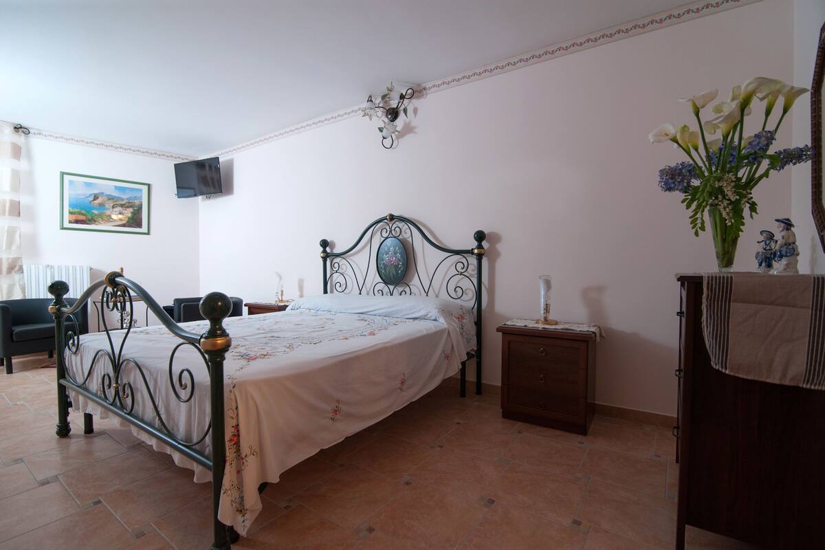 A bedroom with a metal bed frame, a bedside table, a dresser, a TV mounted on the wall, and a painting on the wall.