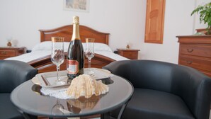 A hotel room with a bed, wooden headboard, a bedside table with a bottle of champagne, two wine glasses, a shell, and a small table with a placemat.