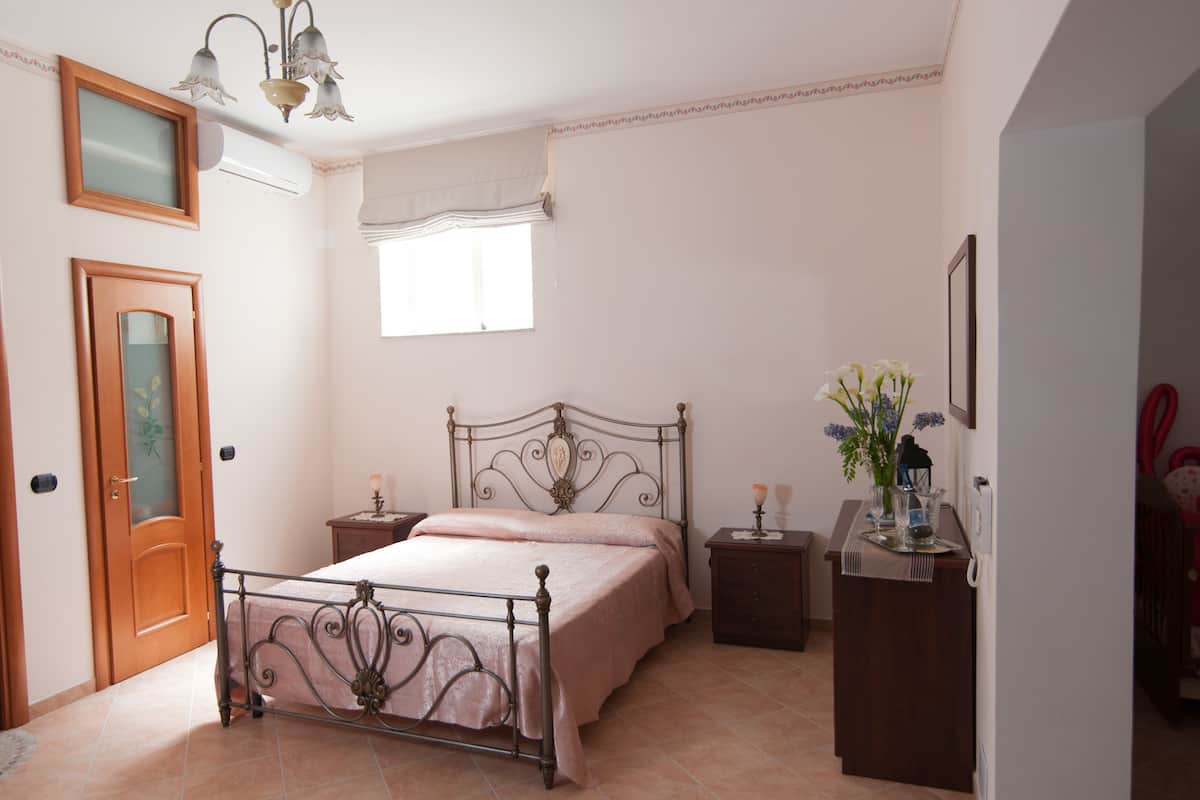 A bedroom with a metal bed frame, a bedside table with a lamp, a nightstand, a window with curtains, and a door with a glass panel.
