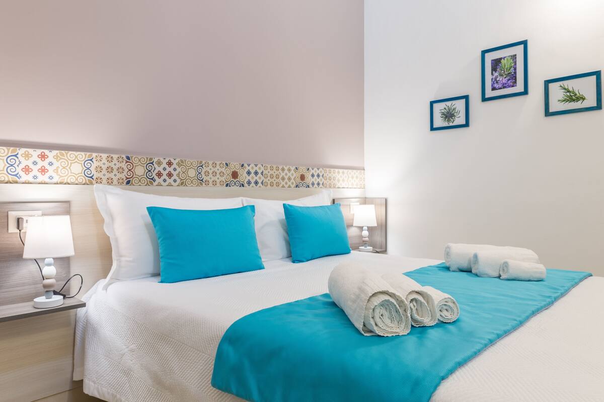 Habitación de hotel con una cama bien hecha, dos lámparas de noche, un cabecero estampado y tres cuadros en la pared.