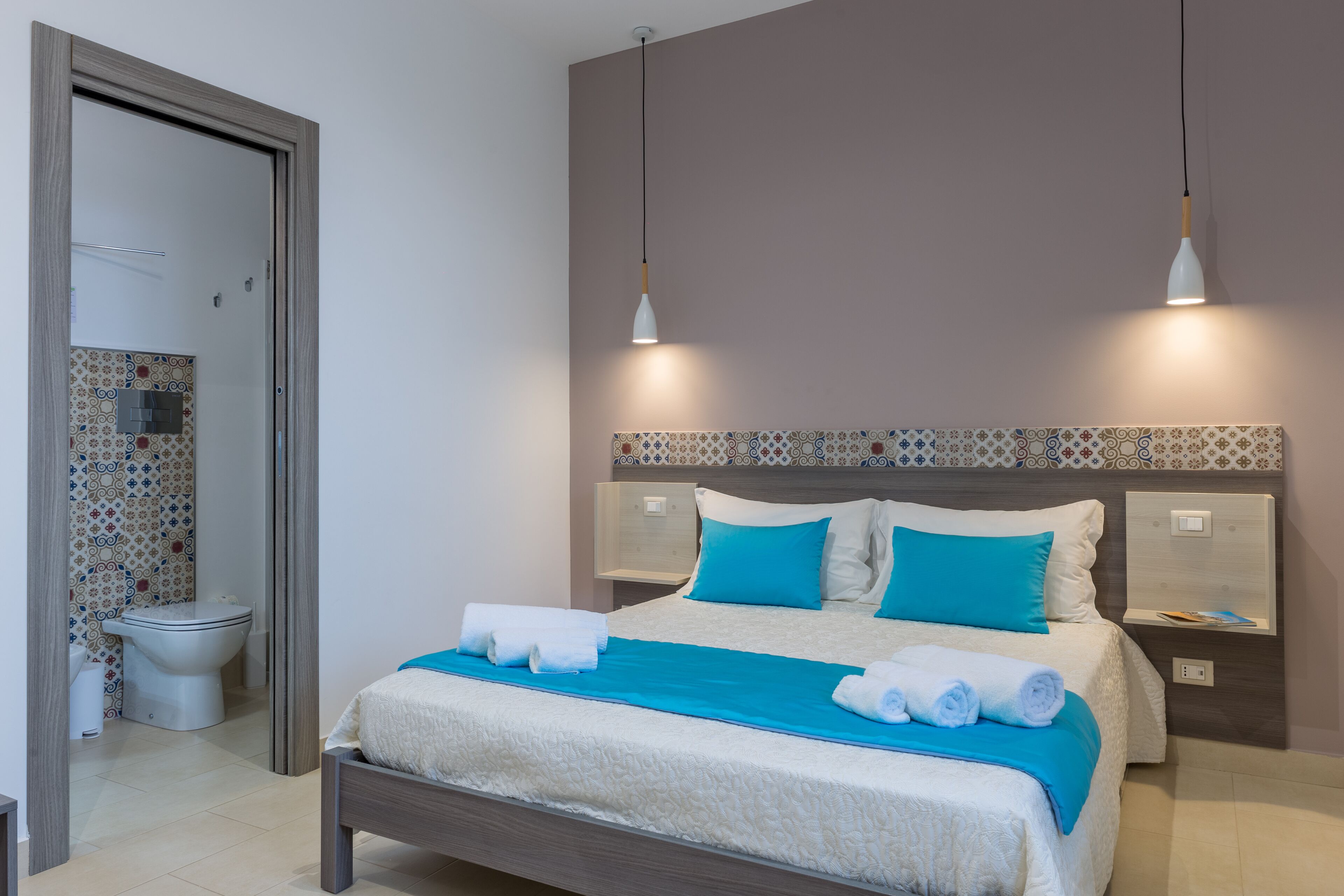A neatly made bed with a blue blanket and white towels, a bedside table with a lamp, and a patterned tile backsplash.