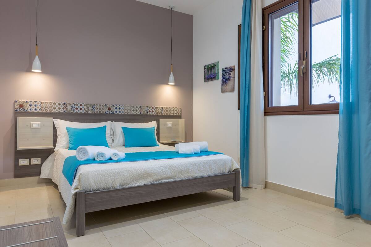 Una cama bien hecha con ropa de cama azul y blanca, dos mesitas de noche, una ventana con cortinas y una pared con un borde de azulejos decorativos.