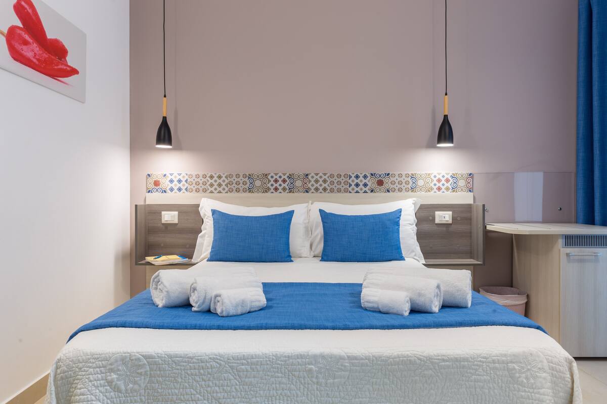 Una cama bien hecha con ropa de cama azul y blanca, dos mesitas de noche con lámparas, un cabecero estampado y una cortina azul.