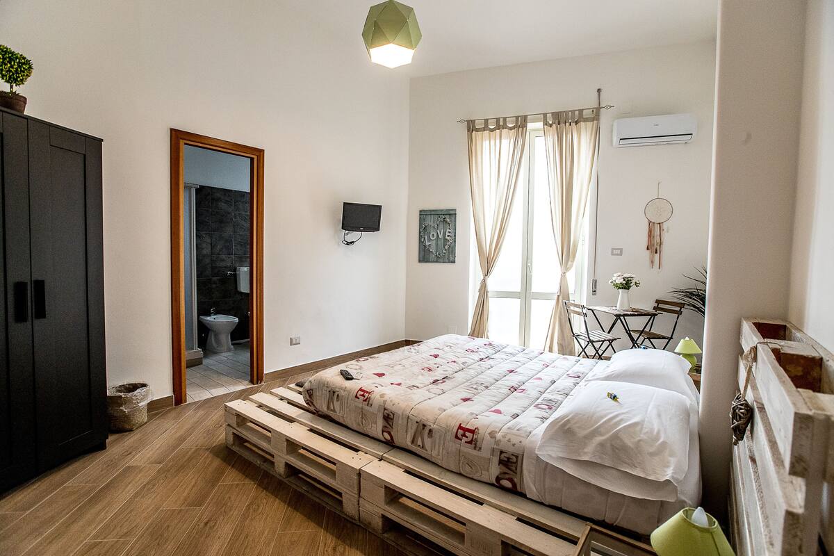 A bedroom with a bed, a television, a window with curtains, a desk with a chair, and a small plant.