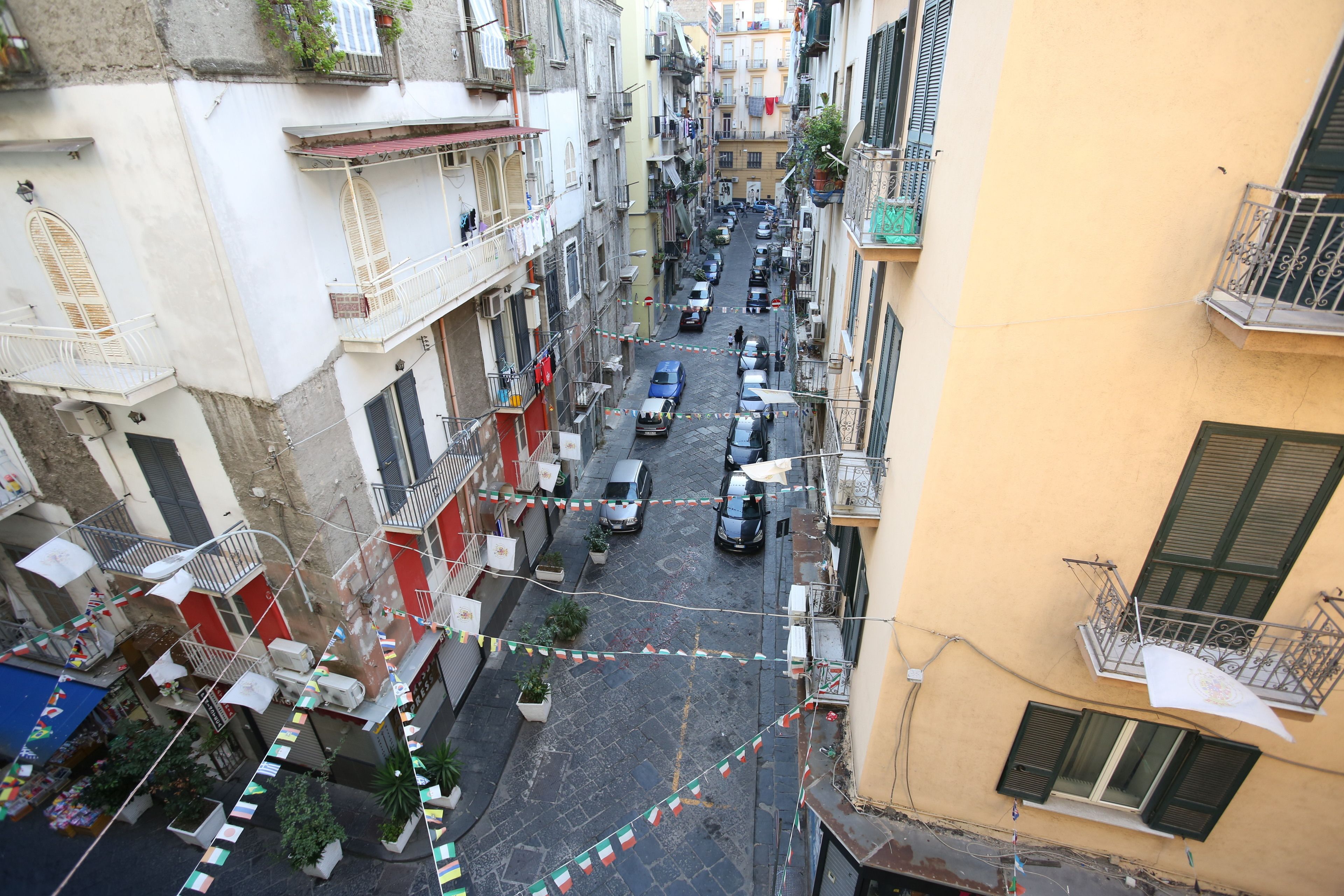 A narrow urban street with buildings on either side, featuring balconies, potted plants, and a variety of parked cars.