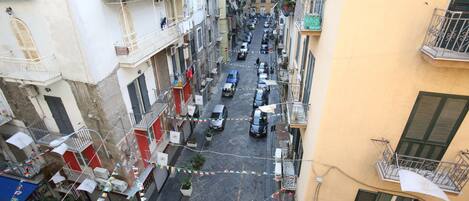 A narrow urban street with buildings on either side, featuring balconies, potted plants, and a variety of parked cars.