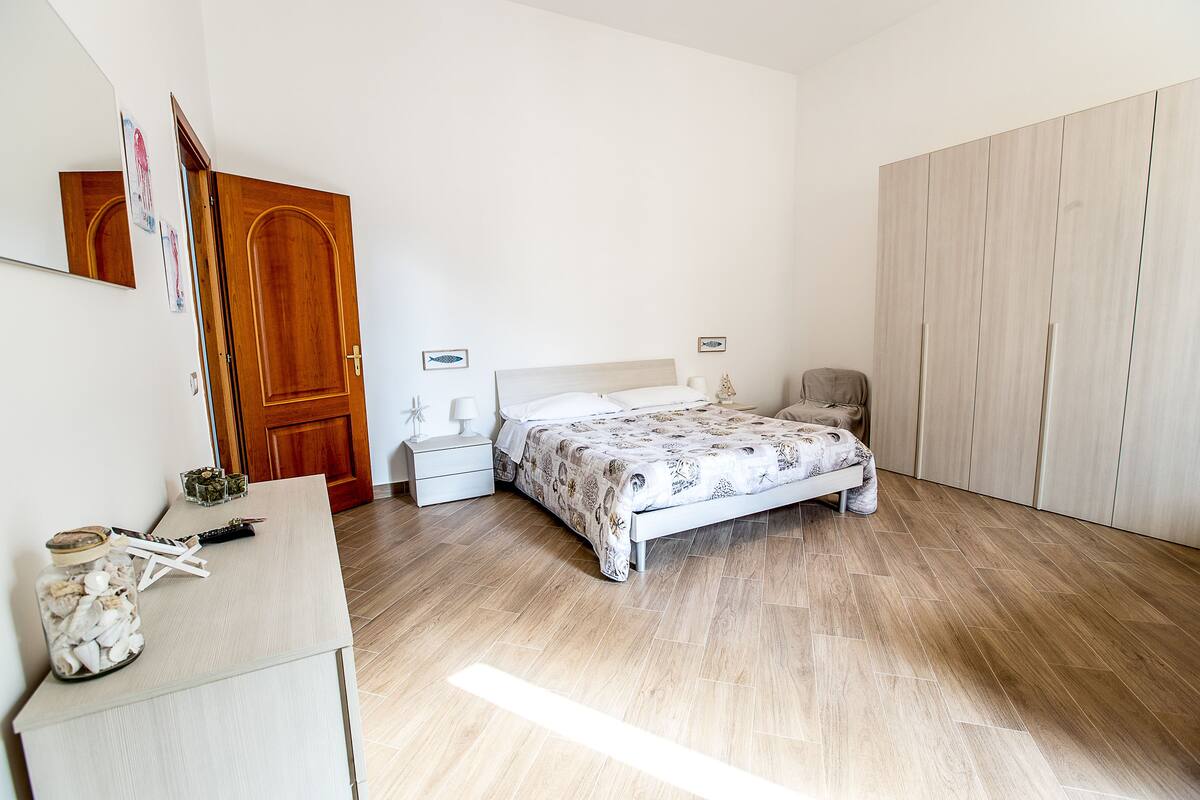 A bedroom with a bed, a dresser, a nightstand, a wooden wardrobe, and a mirror on the wall.