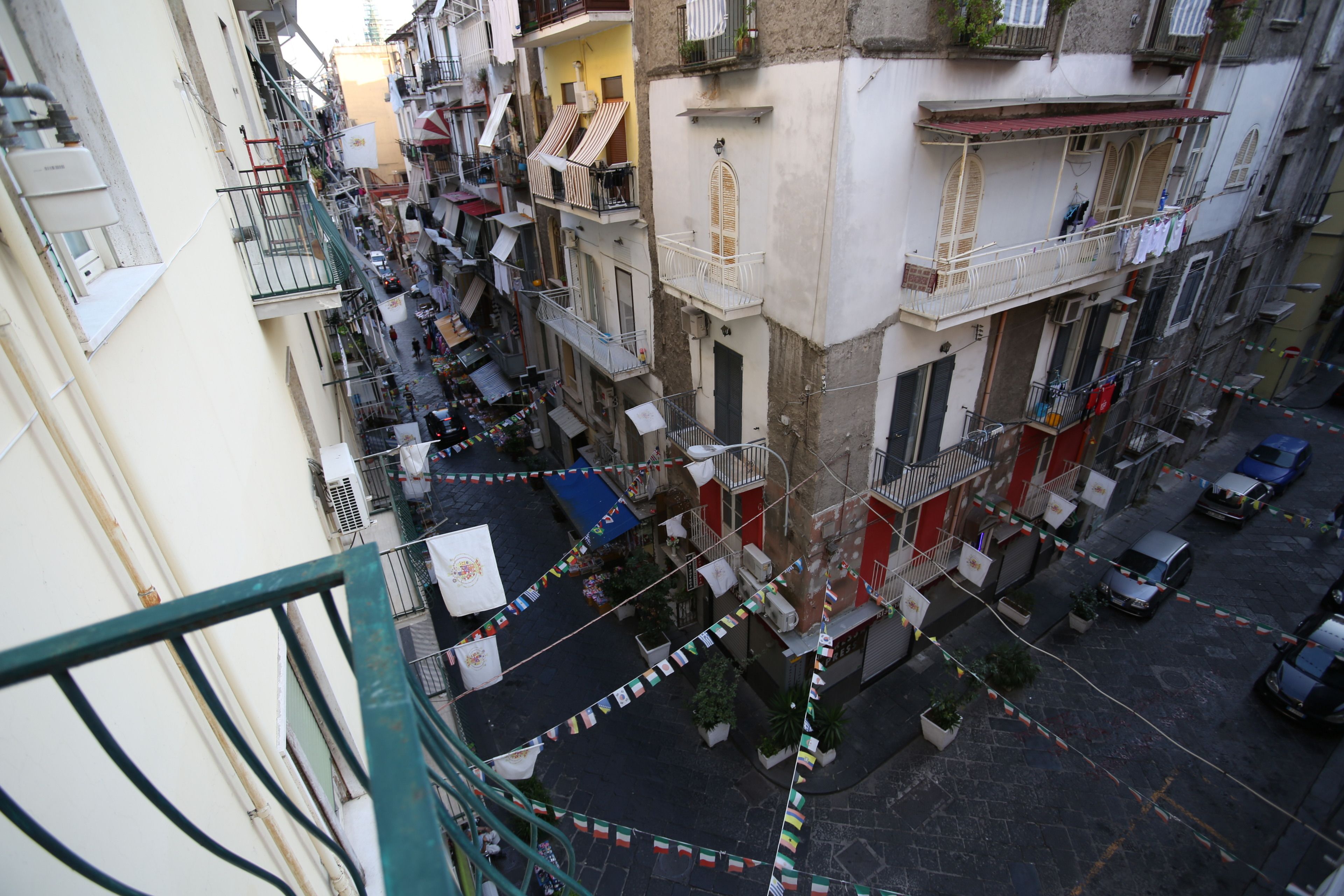 A narrow street with buildings on either side, balconies with railings, and a few parked cars.