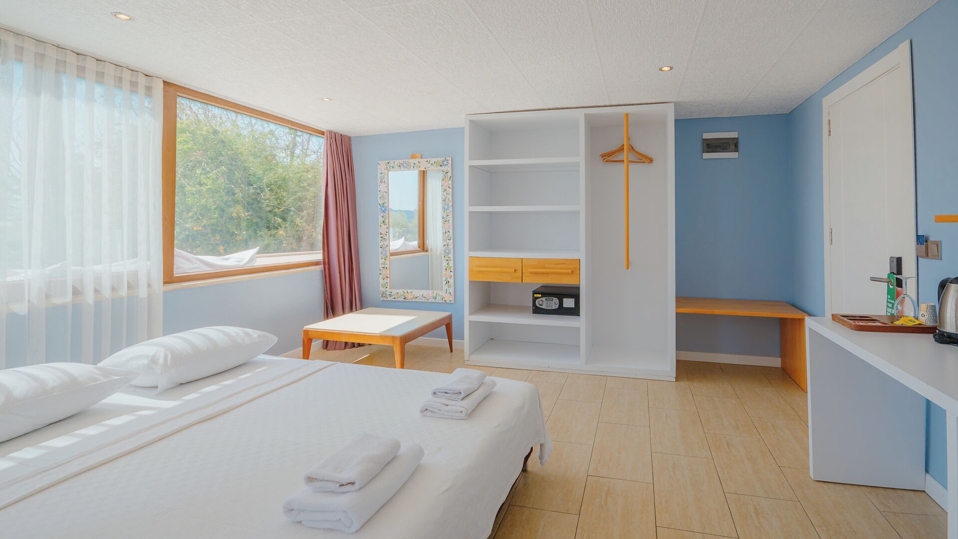 A hotel room with a bed, a bench, a wardrobe, and a desk with a kettle.