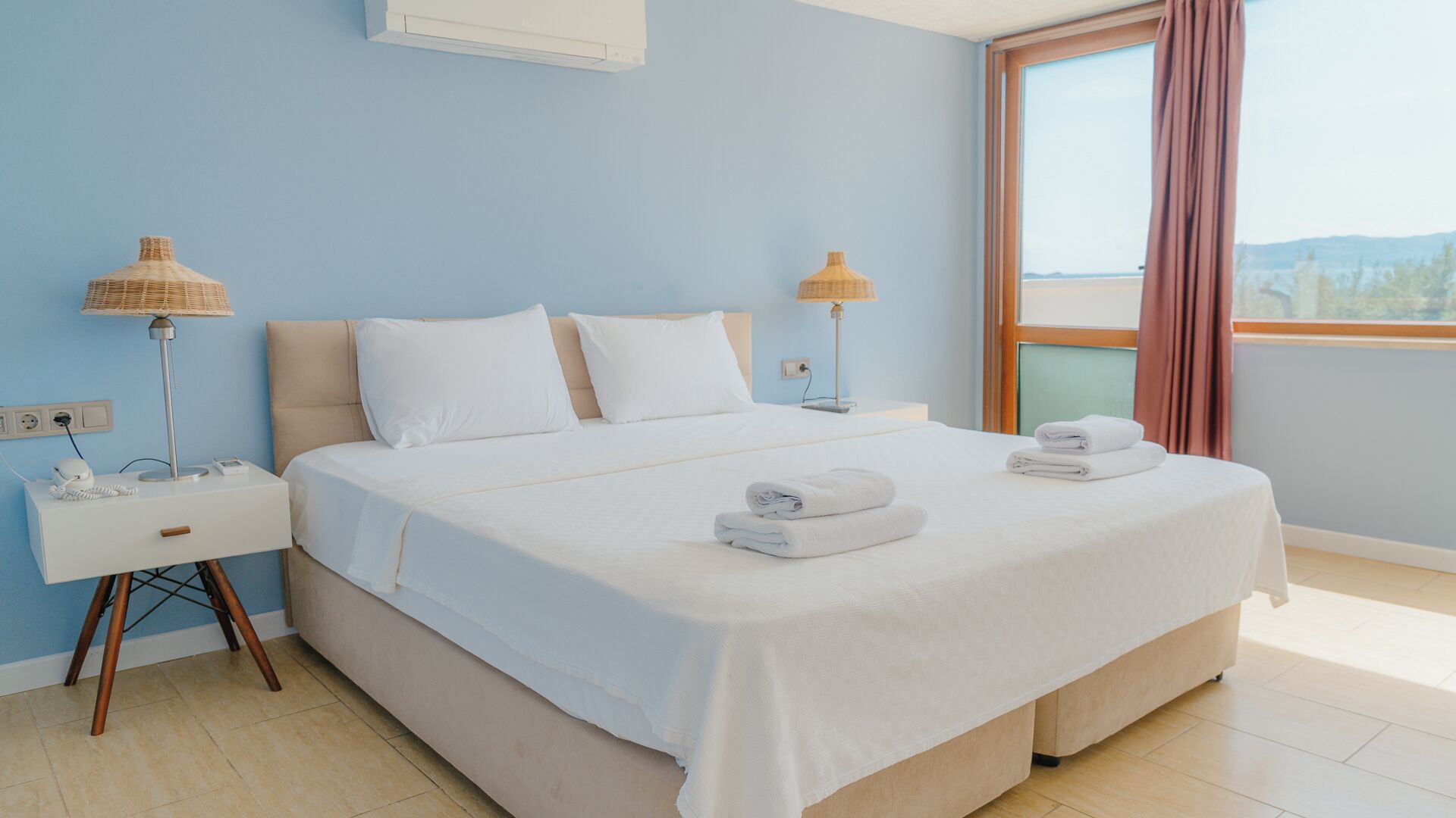 A neatly made bed with white linens and two pillows, a bedside table with a lamp, and a window with curtains.