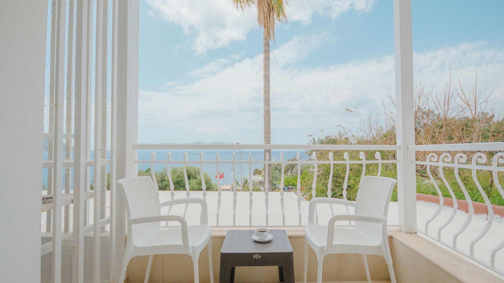 Een balkon met twee witte stoelen, een tafeltje en uitzicht op de zee en palmbomen.