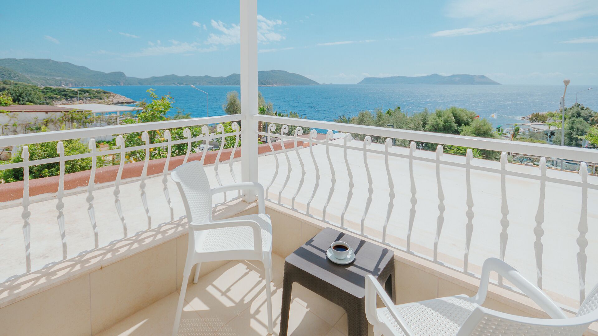 A balcony with white chairs and a small table, overlooking a coastal view with mountains in the distance.