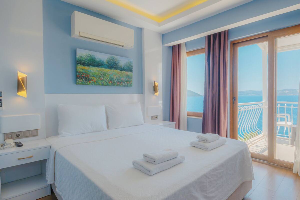 A hotel room with a large bed, a painting, an air conditioning unit, and a balcony with a view of the sea.