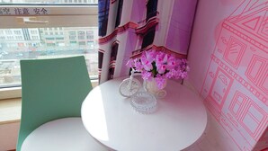 Une pièce avec un fauteuil vert, une table blanche avec un plateau en verre et une fenêtre donnant sur des immeubles et une rue.