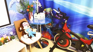 Une pièce comprenant un vélo d’appartement rouge, une petite table sur laquelle est posé un jouet en peluche, un vase avec des fleurs et un mur orné d’une fresque sur le thème de la mer.