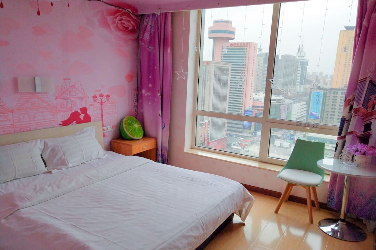 Quarto com cama, mesa de cabeceira, cadeira, pequena mesa e janela com vista para a cidade.