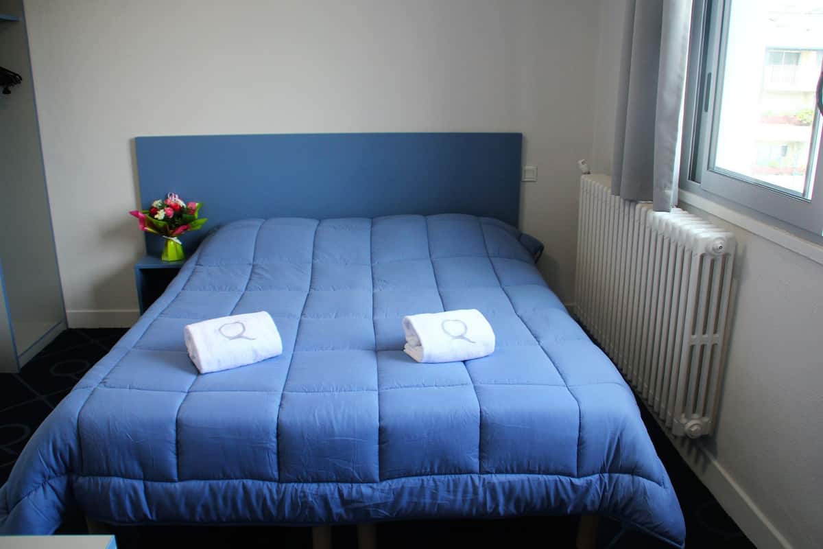 Quarto com colcha azul, duas toalhas com logotipo, um vaso com flores e uma janela com cortinas.