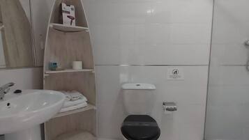 Un baño con lavamanos, inodoro y ducha blancos. Hay un estante con artículos de aseo y un espejo sobre el lavamanos.