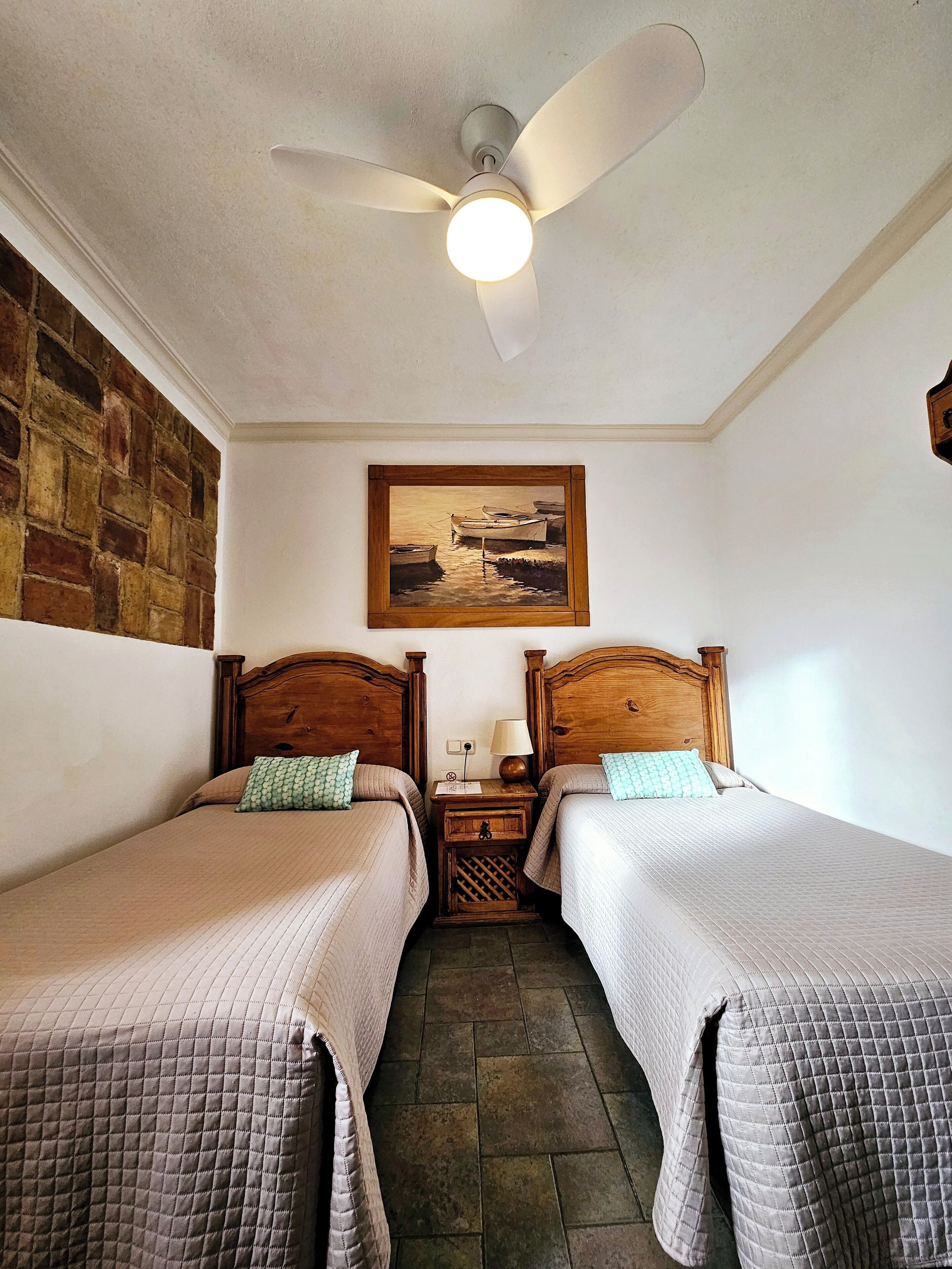 A hotel room with two single beds, a wooden headboard, a nightstand with a lamp, a painting on the wall, and a ceiling fan.