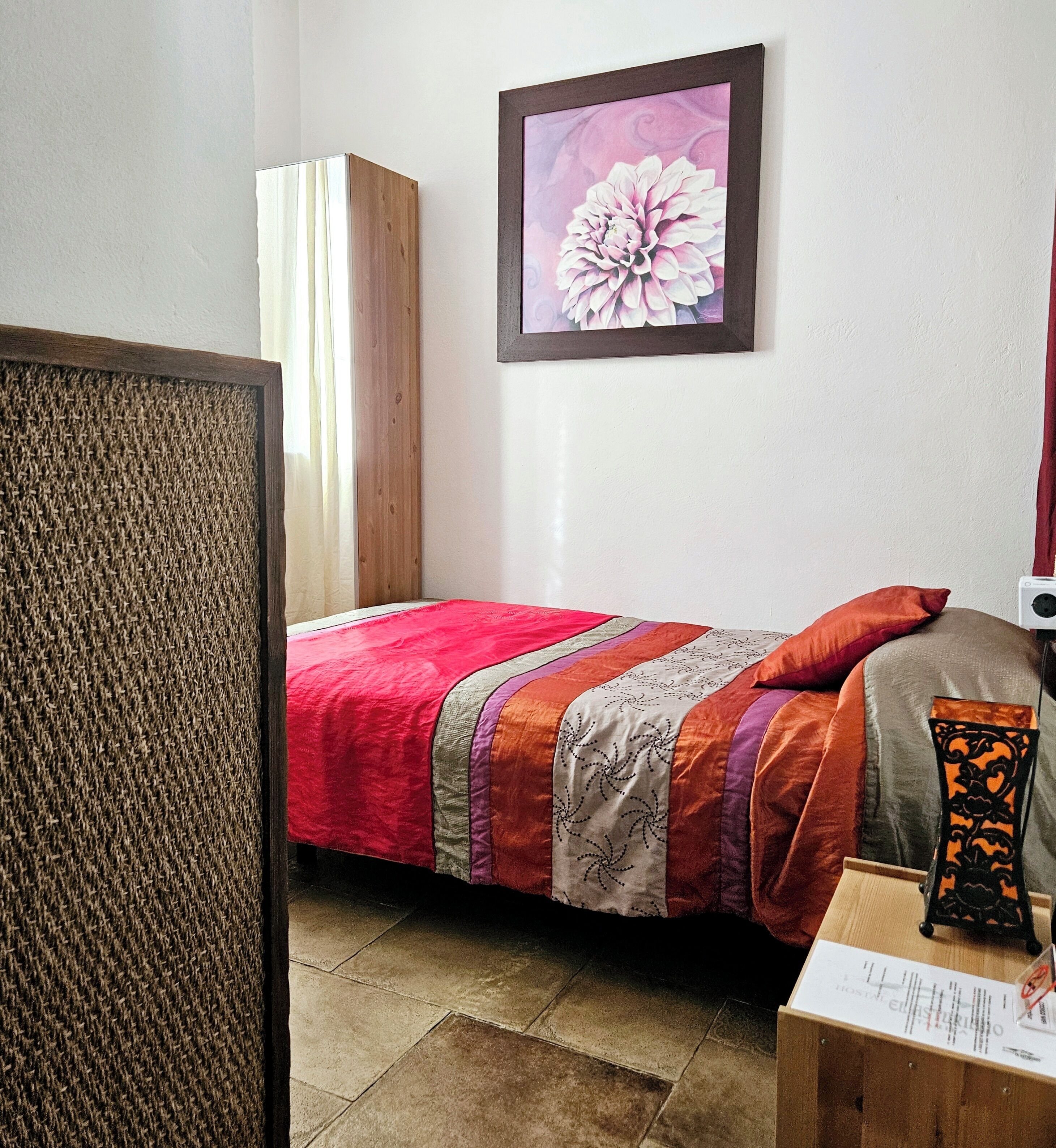 A bedroom with a bed, a wooden headboard, a framed floral artwork, and a bedside table with a lamp.