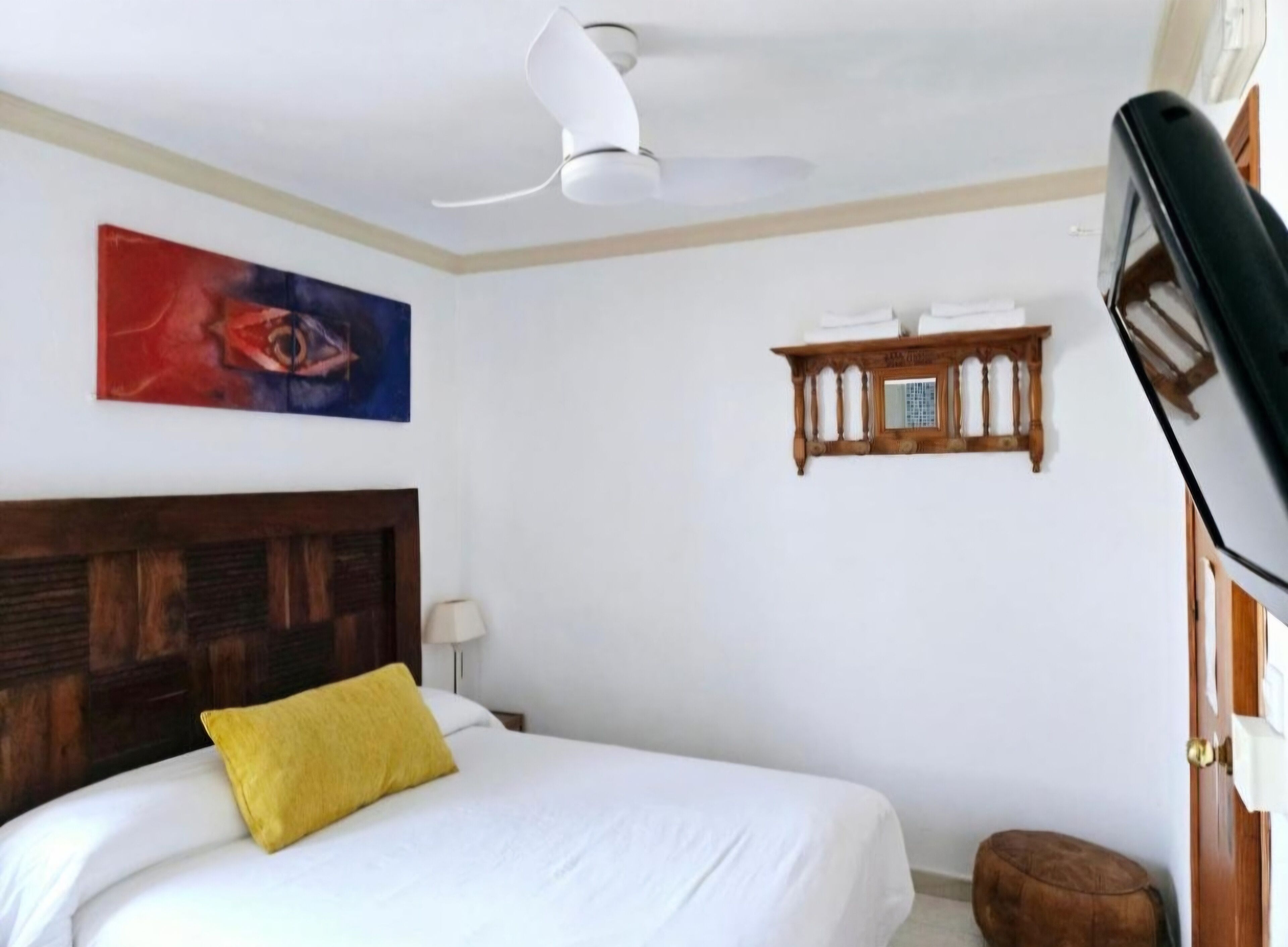 Een hotelkamer met een bed, een plafondventilator, een schilderij, een houten plank met toiletartikelen, en een bruin voetenbankje.