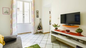 A living room with a grey sofa, a television on a white stand, a small Christmas tree, and a window with curtains. - B&B Dei Mercanti (Salerno)