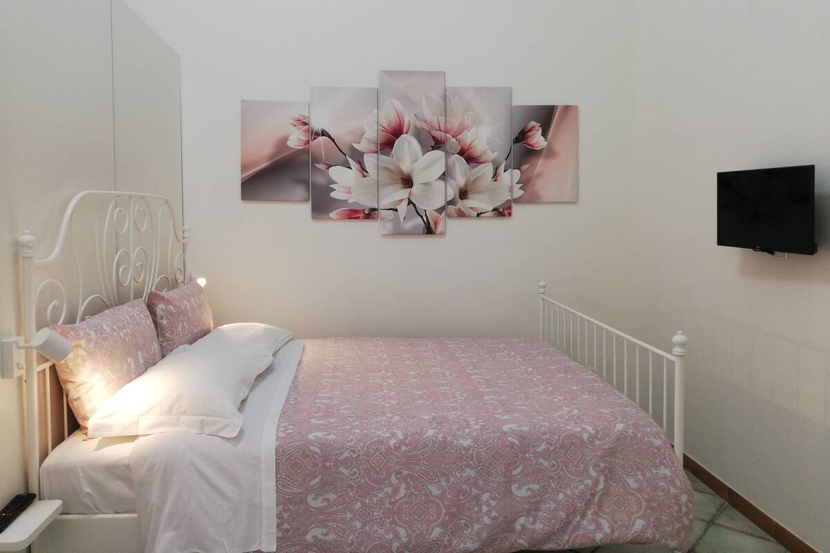 Ein Schlafzimmer mit einem Bett, Kissen, einem an der Wand befestigten Fernseher und einem Blumenbild über dem Bett.
