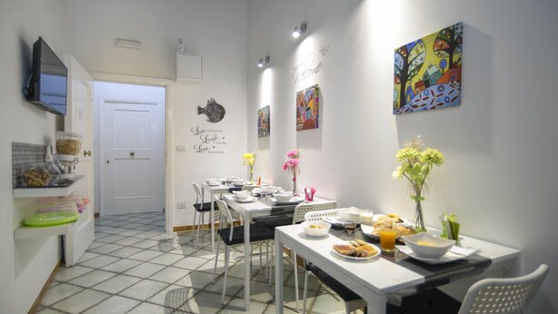 A dining area with white tables and chairs, a television mounted on the wall, and artwork on the walls. - B&B Dei Mercanti (Salerno)
