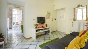 A living room with a sofa, a TV on a white stand, a door, and a window with curtains. - B&B Dei Mercanti (Salerno)