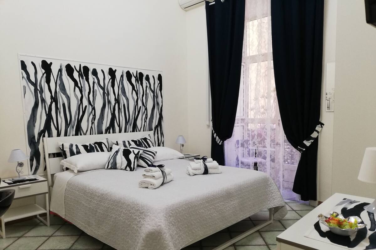 Ein ordentlich bezogenes Bett mit einem zebra-gestreiften Kissen, ein Nachttisch mit einer Lampe und ein Fenster mit transparenten Vorhängen.