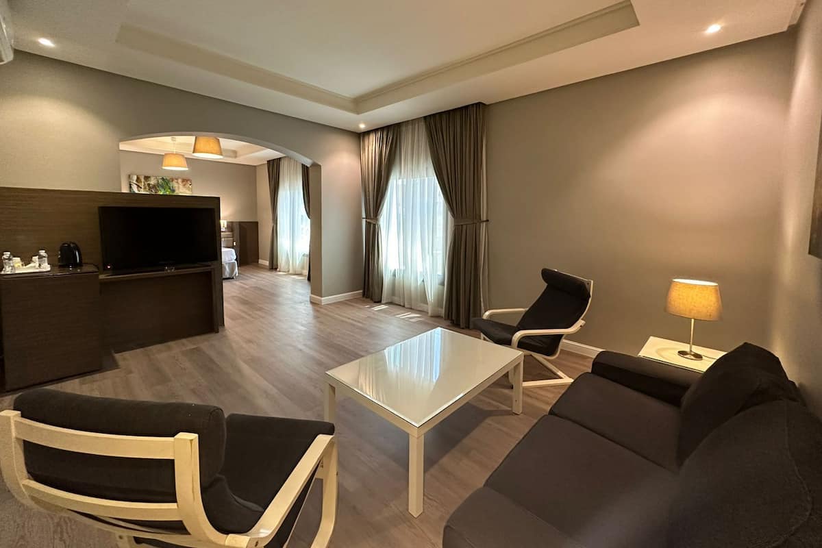 A modern hotel room with a living area, a sofa, a coffee table, a TV, and a desk with a lamp.