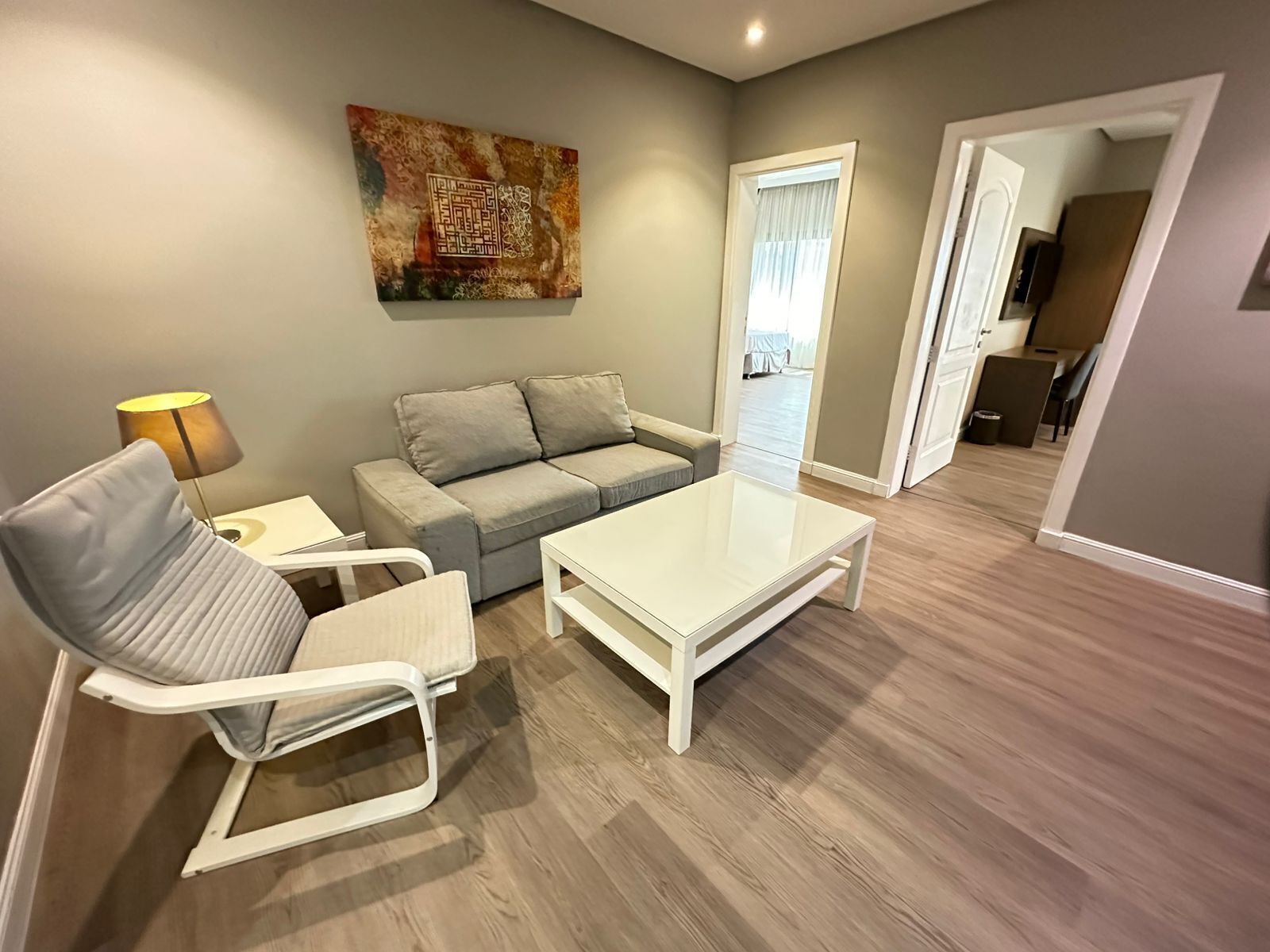 A modern living room with a sofa, a coffee table, and a chair. There is a painting on the wall and a window with curtains.