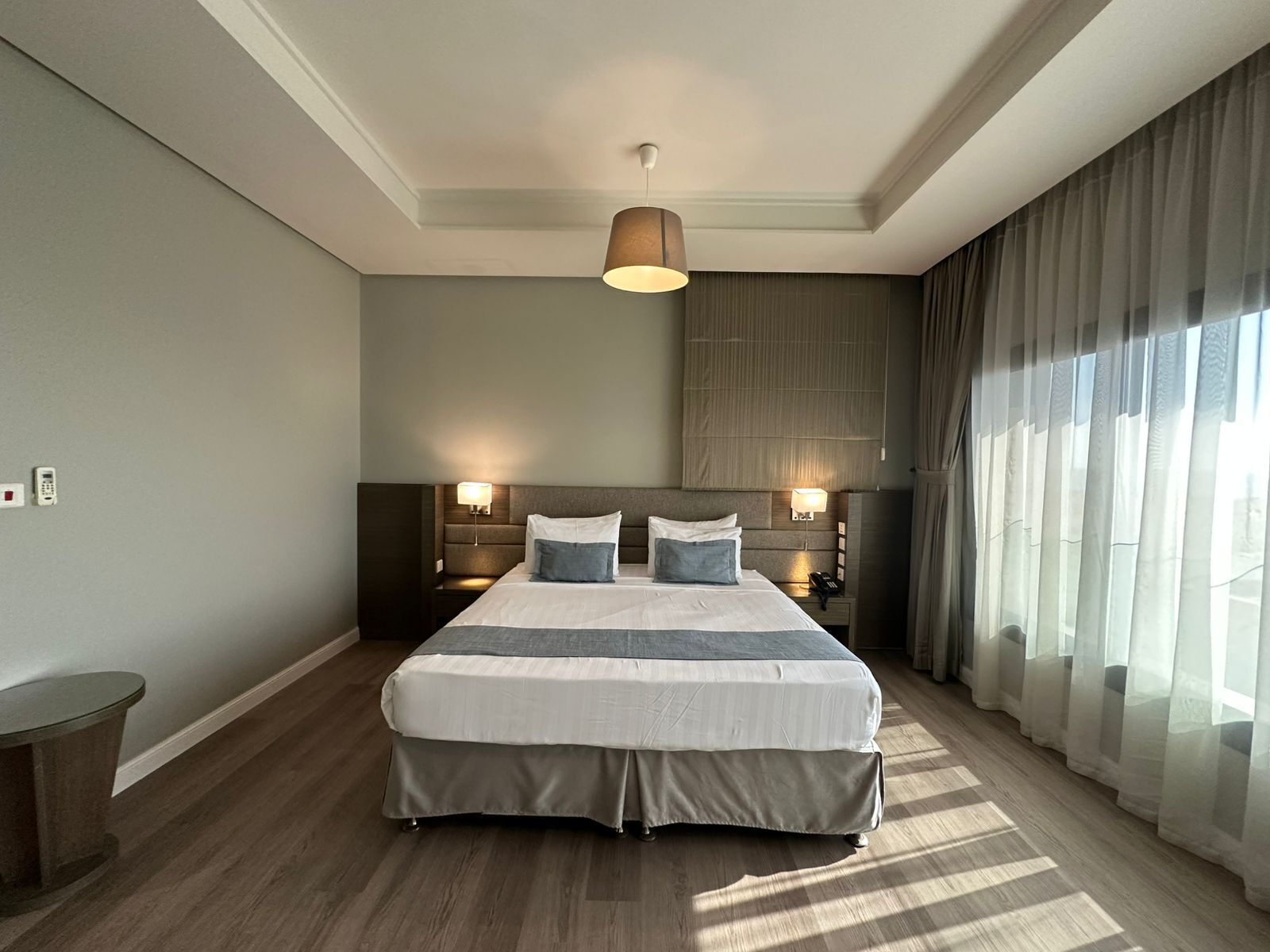 A modern hotel room with a large bed, two bedside lamps, a small table, and a large window with curtains.