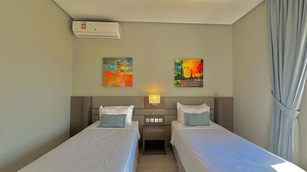 Quarto de hotel com duas camas de solteiro, mesa de cabeceira com luminária e duas pinturas abstratas na parede.