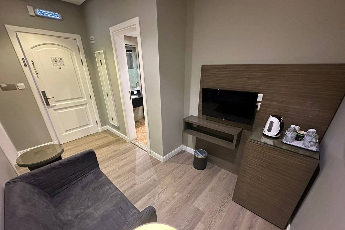 A compact hotel room with a sofa, a TV, a small table with a kettle, and a trash bin.